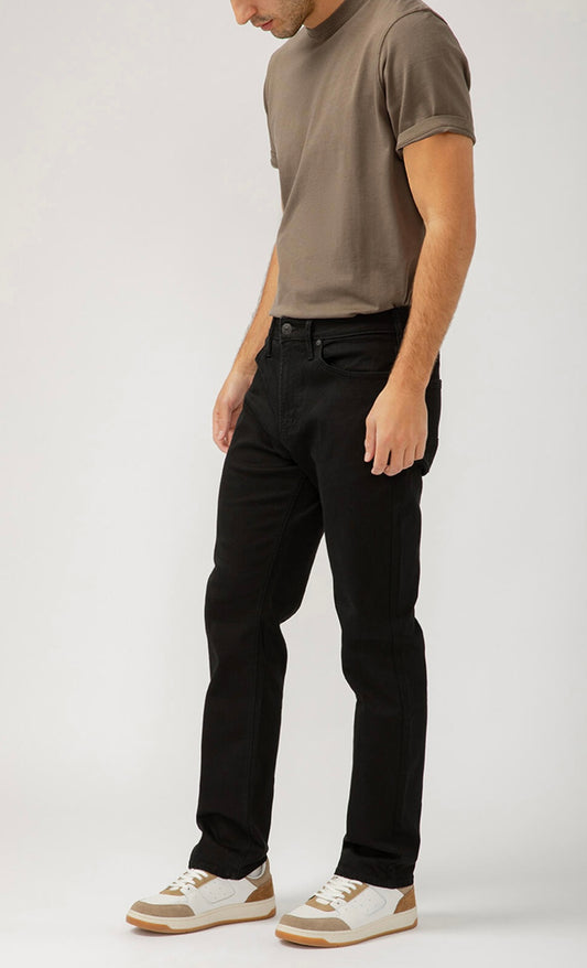 A man stands against a plain background wearing a taupe t-shirt, Machray Athletic Fit Straight Leg black jeans from Silver Jeans Co., and white sneakers with tan accents. His head is slightly turned down and his arms are relaxed at his sides.