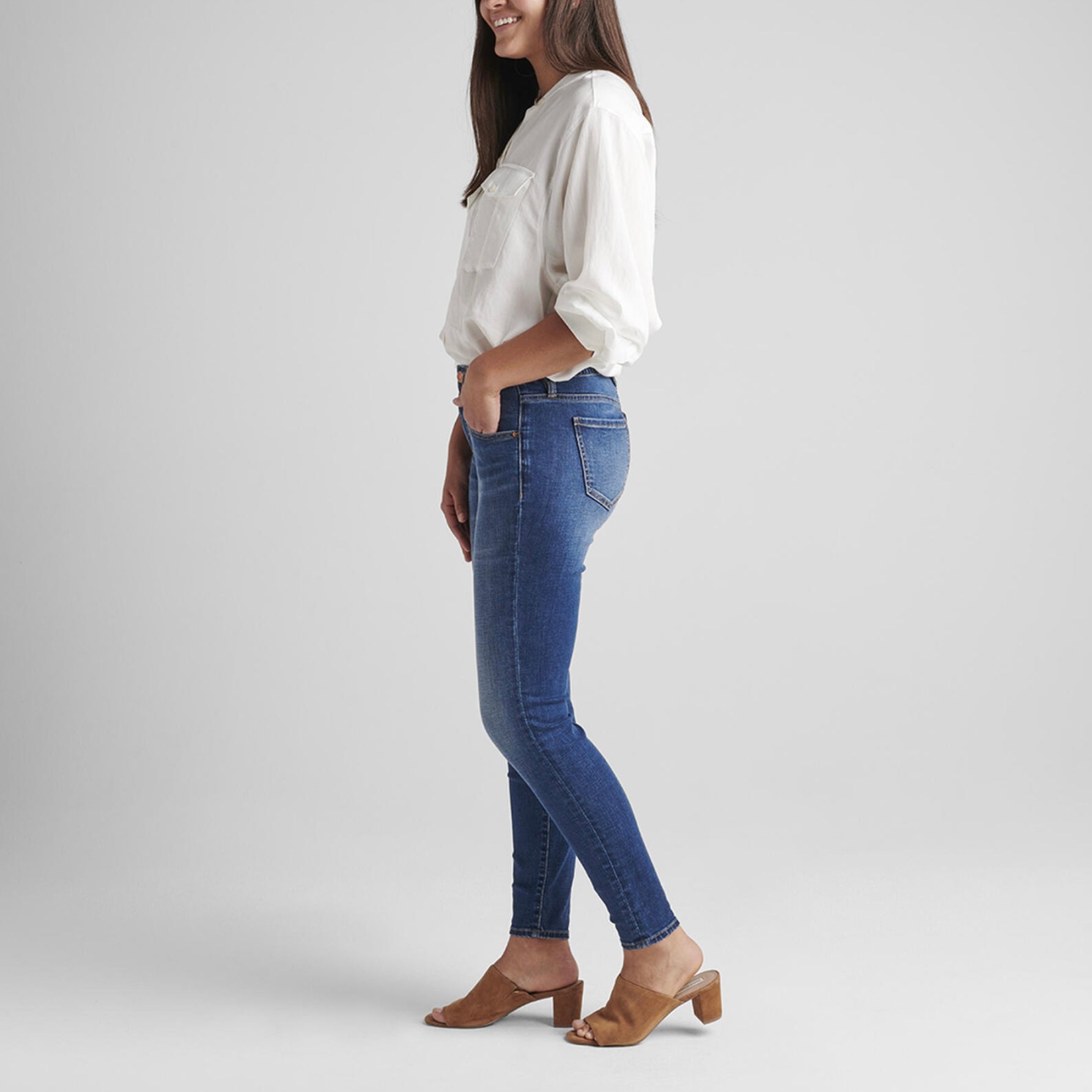 A person stands against a plain background, wearing a white, long-sleeved shirt with front pockets, tucked into Cecilia Mid Rise Skinny Jeans featuring Best Kept Secret technology by JAG. They are also wearing brown, open-toe heeled sandals. One hand is tucked into the jeans pocket while the other hangs at their side.