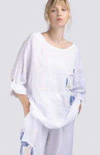 A woman stands with her hands in her pockets, wearing the Paolo Tricot Top 100% Linen DM61163 and matching white pants with blue and beige embroidered patches, set against a light gray background.
