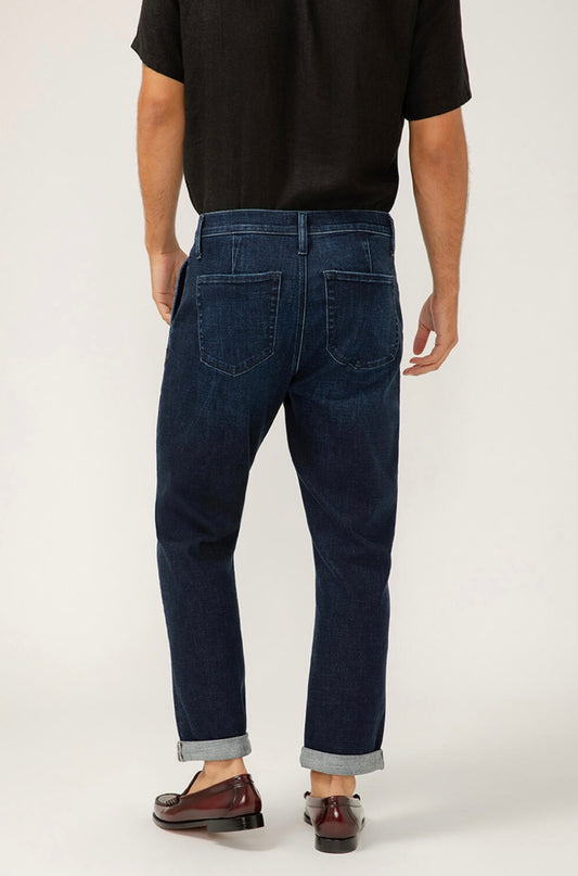 A person in a black shirt, Silver Jeans Co. Trouser Leg Jeans (SKU# M23911BTV445) in dark indigo with cuffed hems, and brown loafers stands facing away from the camera against a plain background.