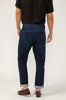 A person in a black shirt, Silver Jeans Co. Trouser Leg Jeans (SKU# M23911BTV445) in dark indigo with cuffed hems, and brown loafers stands facing away from the camera against a plain background.