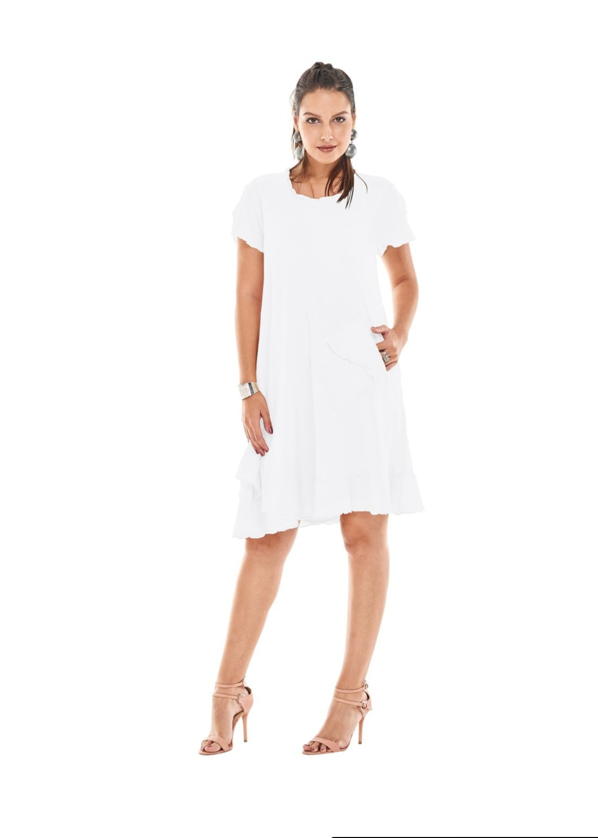 A woman with dark hair is posing in the LA DRESS SNOW by Oh My Gauze, a short-sleeve, white cotton gauze dress featuring cutout shoulder details that reaches just above her knees. She is wearing beige high-heeled sandals and has one hand in her dress pocket. The background is white.