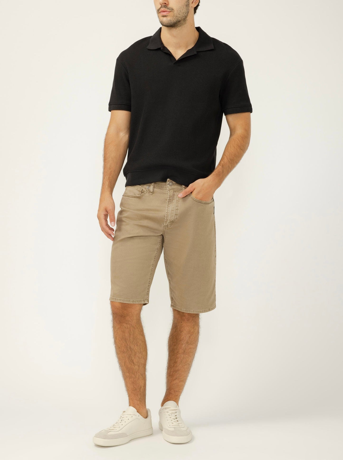 A man stands against a plain background wearing a black polo shirt, Silver Jeans Co. Zac Relaxed Fit Essential Twill Shorts (M52402CBT675), and white sneakers. One hand is in his pocket, and his head is slightly turned to the side.