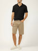 A man stands against a plain background wearing a black polo shirt, Silver Jeans Co. Zac Relaxed Fit Essential Twill Shorts (M52402CBT675), and white sneakers. One hand is in his pocket, and his head is slightly turned to the side.
