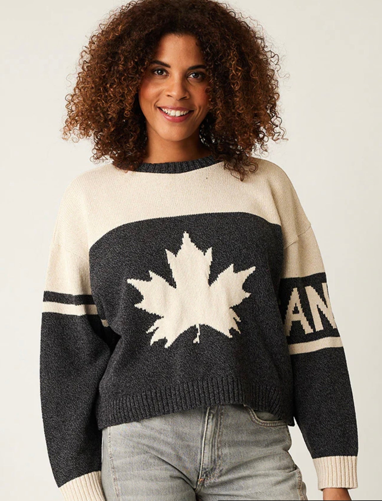 A person with curly hair smiles while wearing a Parkhurst CANADA SHORTY SWEATER 87322, featuring a large maple leaf design on the front. This eco-friendly black and white sweater pairs perfectly with their light-colored jeans.