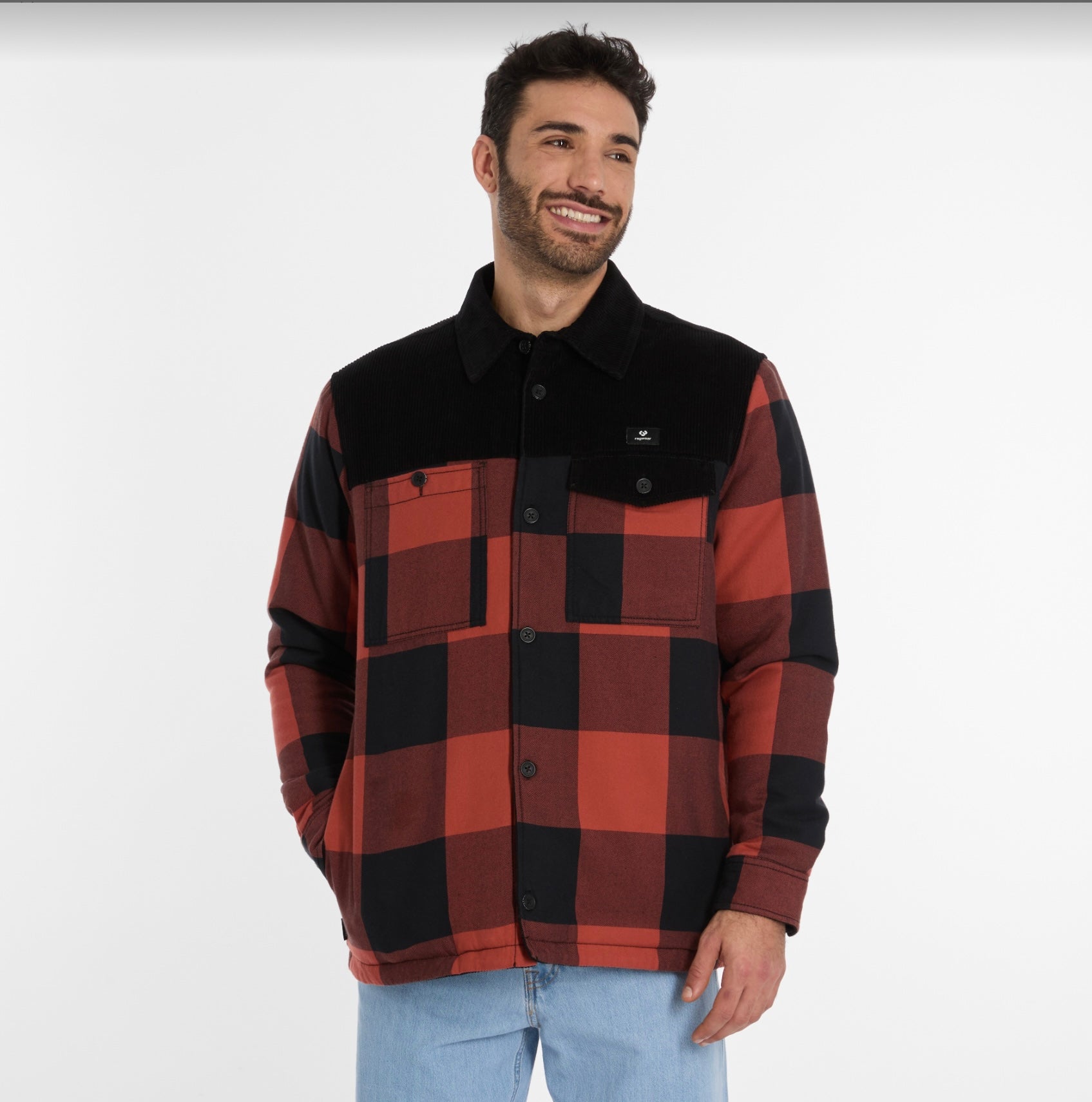 A man with short dark hair and a beard is wearing a CHECON Men's Jacket by RAGWEAR in red and black plaid, featuring a black collar and pocket flaps. He pairs it with light blue jeans and smiles against a plain white background.