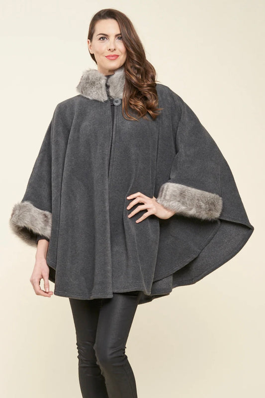 A woman is wearing the Desmona Faux Fur Trim Fleece Cape 22606 by Parkhurst, featuring a dark gray hue with luxurious faux fur trim around the collar and sleeves. Her long brown hair cascades down as she stands against a neutral background, smiling slightly and posing with one hand on her hip, embodying elegance that's perfect for any occasion.