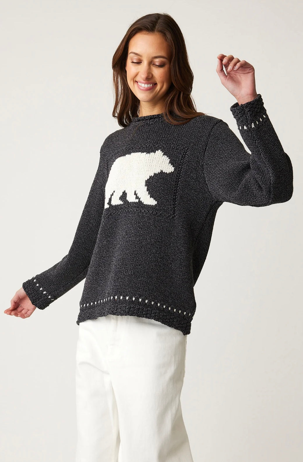 A person is smiling and looking down while wearing a Parkhurst Cotton Country Laurentian Bear Pullover in dark gray, featuring a white polar bear pattern. Their long hair complements their white pants, and the plain background highlights both the relaxed fit of the pullover and their joyful expression.