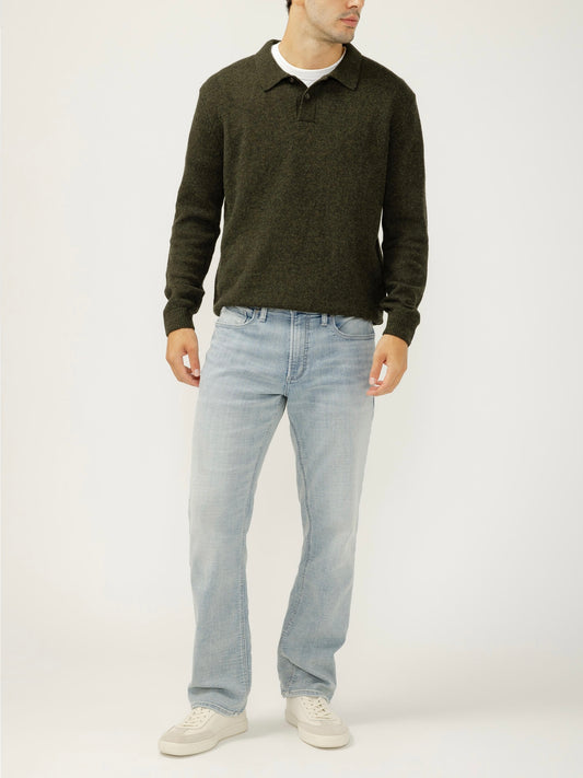 A man stands against a plain background wearing a dark green long-sleeve knit collared shirt, Silver Jeans Co. Grayson Classic Fit Straight Leg Jeans (M23425EWK112) in medium indigo, and white sneakers. His hands are at his sides, face partially out of frame.