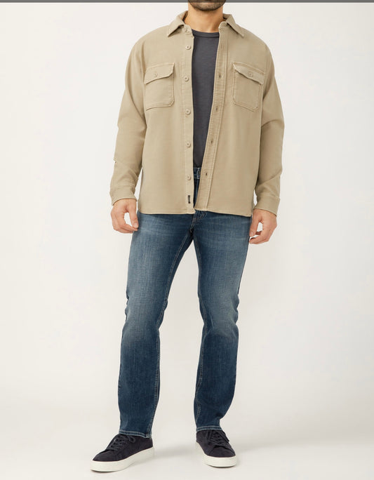 A man, with his head out of frame, is wearing a Silver Jeans Co. Button Up Knit Flex Shirt MJ0035KTS668 SILVER over a dark gray tee, paired with blue knit flex denim jeans and black sneakers against a plain background.