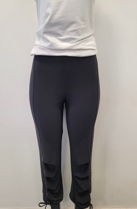 A person models Bali Corp’s Black Leggings 6279-Bali Corp, featuring decorative button and tie details near the ankles, paired with a white t-shirt, against a plain light-colored wall. Made in Canada.