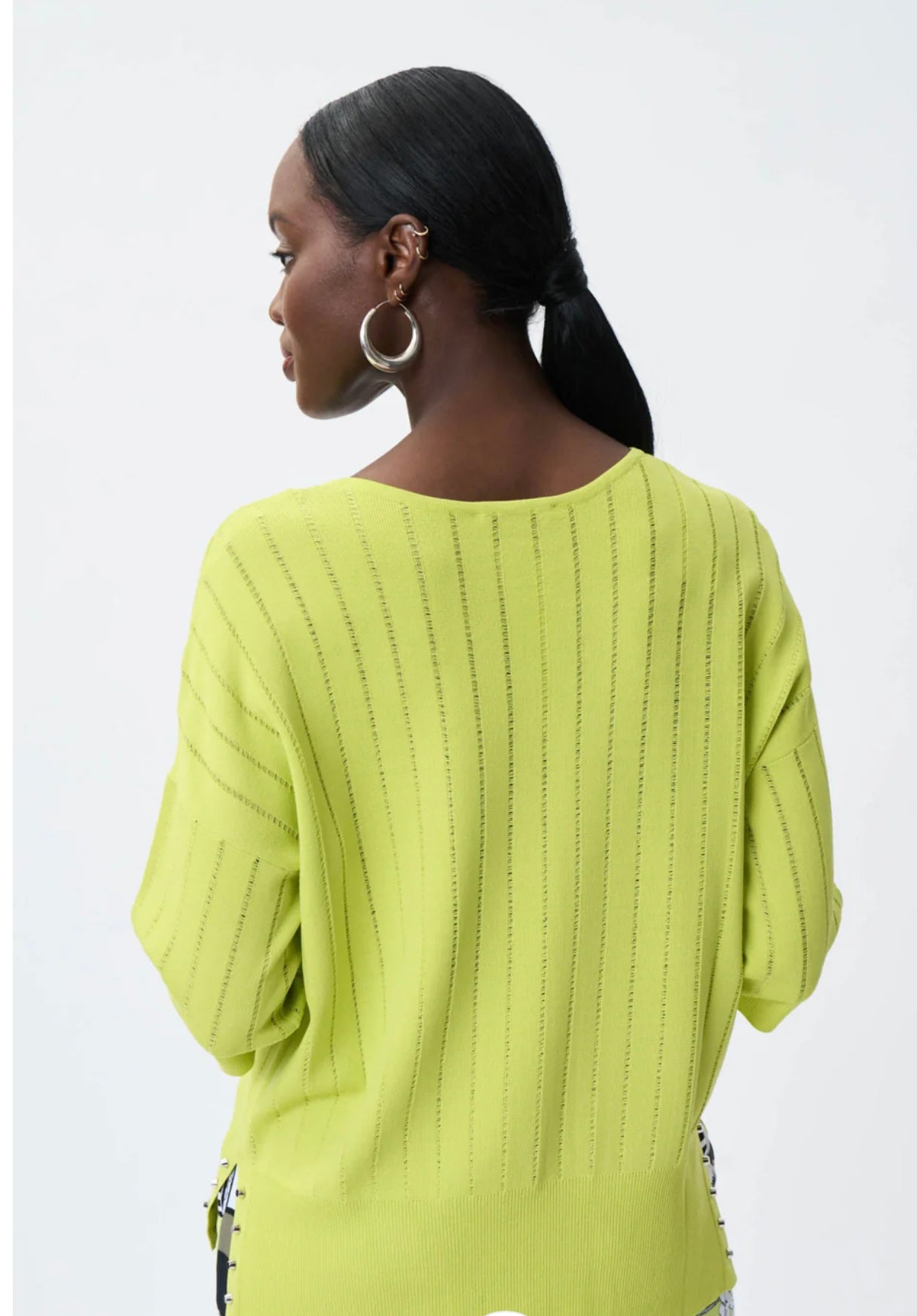 A stylish person with dark skin and short hair is wearing a Green Striped Shirt from Joseph Ribkoff over a white shirt. They have gold hoop earrings and are touching their neck with one hand, exuding an elegant vibe against the plain white background.