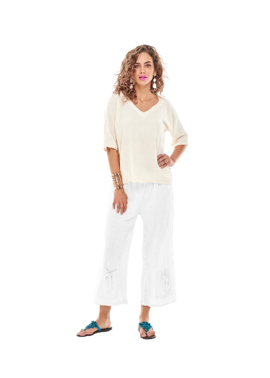 A woman stands against a white background, wearing a light beige V-neck top and the SAMMY PANTS SNOW from Oh My Gauze, featuring subtle embroidery and tie pocket detail. Her turquoise sandals and gold bracelets complement the outfit, while her curly hair frames her neutral expression as she looks directly at the camera.