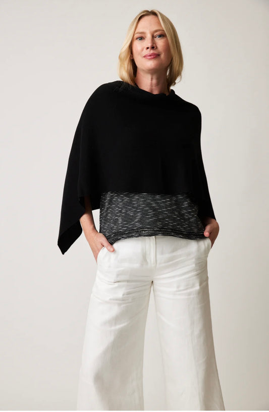 A person with shoulder-length hair is wrapped in a Covi Wrap STYLE 22081 by Parkhurst, a versatile black poncho with a striped hem. They have paired it perfectly with white wide-leg pants and have one hand in their pocket, exuding confidence against a plain background.