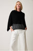 A person with shoulder-length hair is wrapped in a Covi Wrap STYLE 22081 by Parkhurst, a versatile black poncho with a striped hem. They have paired it perfectly with white wide-leg pants and have one hand in their pocket, exuding confidence against a plain background.