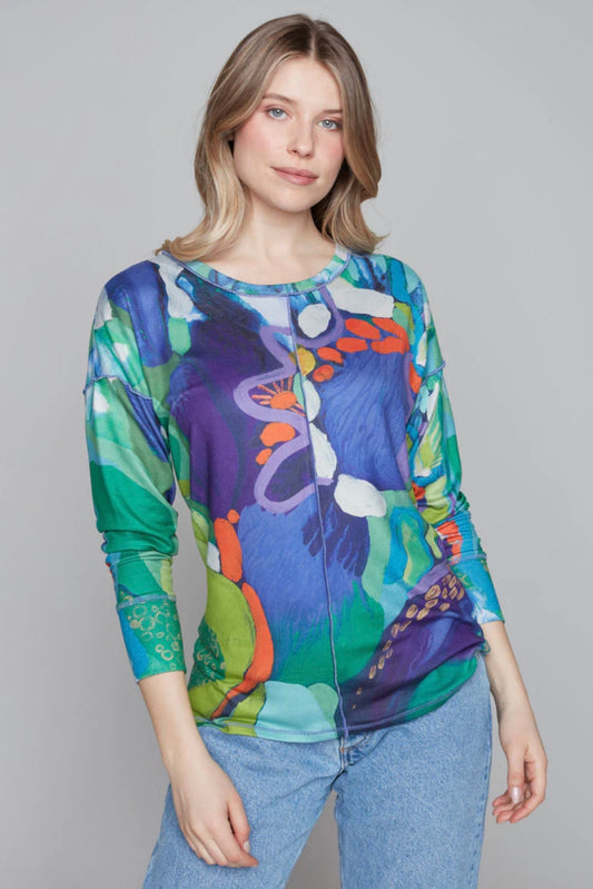 A person with wavy, shoulder-length hair stands against a plain gray background wearing Knit Top 91736 by Claire Desjardins, featuring a long-sleeved, jewel-toned abstract pattern reminiscent of Claire Desjardins' artwork, paired with light blue jeans. They are posing with one hand resting on their hip and the other arm relaxed by their side.