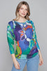 A person with wavy, shoulder-length hair stands against a plain gray background wearing Knit Top 91736 by Claire Desjardins, featuring a long-sleeved, jewel-toned abstract pattern reminiscent of Claire Desjardins' artwork, paired with light blue jeans. They are posing with one hand resting on their hip and the other arm relaxed by their side.