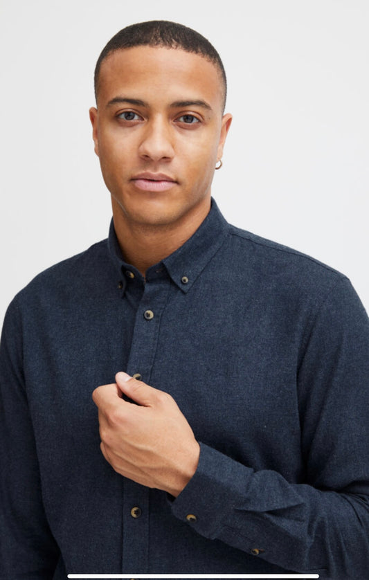 A person with short hair wears a dark BHBURLEY Shirt -BLEND by BLEND, standing against a plain light background, looking at the camera and holding the cotton button-up collar with their left hand.