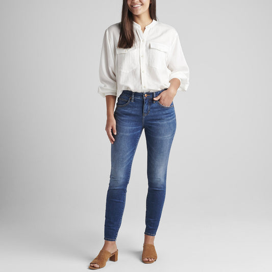 A person stands against a plain background, wearing a white, long-sleeved shirt with front pockets, tucked into Cecilia Mid Rise Skinny Jeans featuring Best Kept Secret technology by JAG. They are also wearing brown, open-toe heeled sandals. One hand is tucked into the jeans pocket while the other hangs at their side.