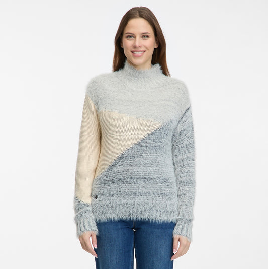 A woman with long brown hair smiles while wearing the RAGWEAR YENOVEFA Sweater, a turtle neck style with diagonal color blocks in light gray, cream, and blue. She stands against a plain white background and pairs it with classic blue jeans.