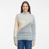 A woman with long brown hair smiles while wearing the RAGWEAR YENOVEFA Sweater, a turtle neck style with diagonal color blocks in light gray, cream, and blue. She stands against a plain white background and pairs it with classic blue jeans.