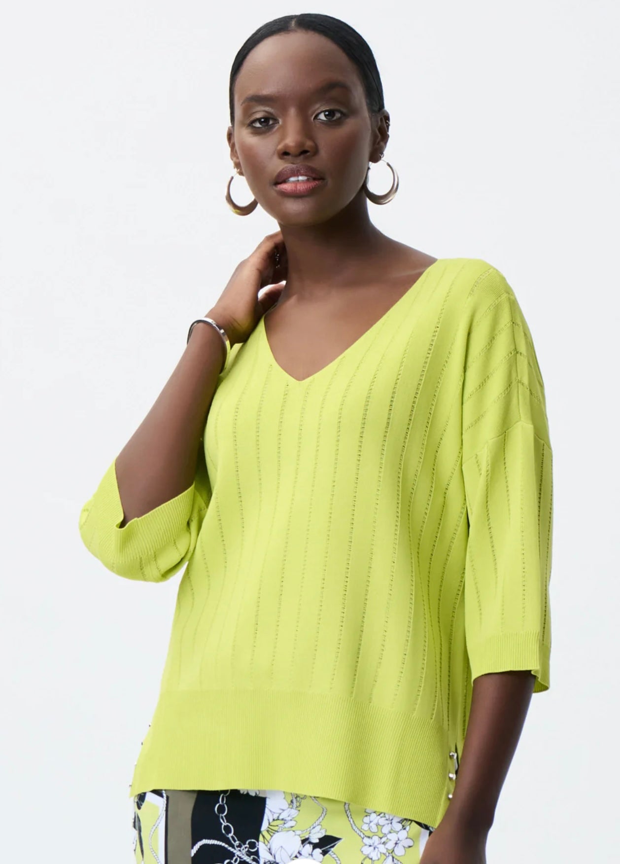 A woman wearing a Green Striped Shirt (231944) by Joseph Ribkoff, paired with floral-patterned pants. She has hoop earrings, and her left hand is gently touching her neck. The background is plain white, making this comfortable and stylish outfit a must-have wardrobe addition.