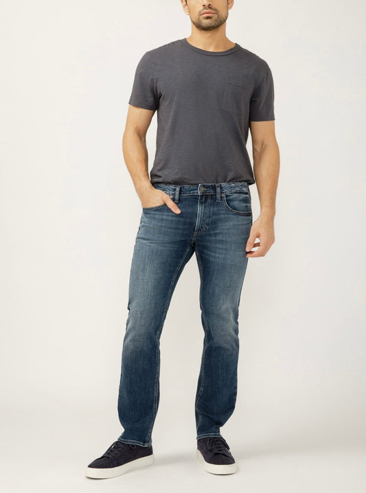 A man poses against a white background wearing a dark gray t-shirt, dark sneakers, and Silver Jeans Co. Allan Slim Fit Slim Straight Leg Jeans M12401SDK361 in a medium-dark indigo Max Flex denim. One hand is in his pocket; his face is partially out of frame.