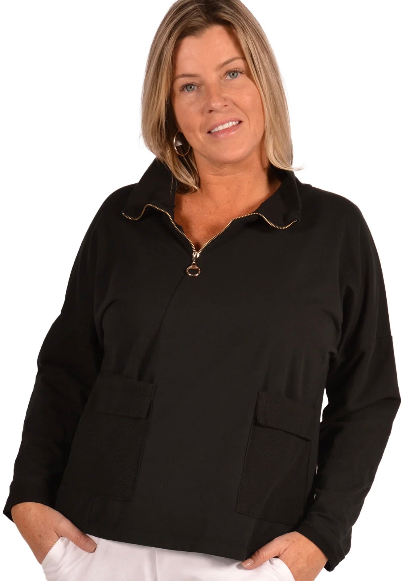 A woman with shoulder-length blonde hair is smiling and wearing the Catherine Lillywhite ITM5093BK Sweater, a black half zip top with two pockets. She stands with her hands in the pockets, facing the camera against a plain white background.