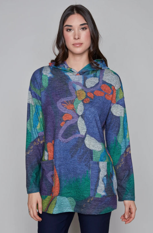 A woman with long dark hair wears a colorful Knit Top 91782 from Claire Desjardins, displaying an abstract pattern in hues of blue, green, orange, and white. She stands against a plain background with her hands relaxed by her sides.