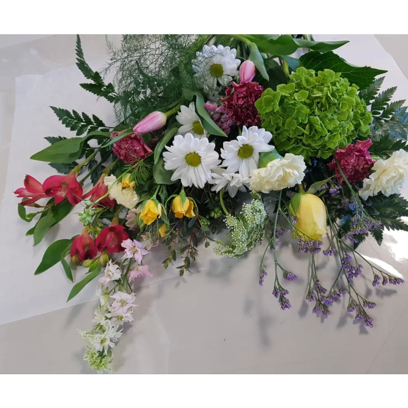 Introducing the Garden Mix by Posies Flowers & Fashion: a vibrant floral arrangement featuring a diverse collection of flowers, including white daisies, pink tulips, yellow bells, red and pink carnations, white roses, purple sprigs, and lush greenery. Artfully arranged in a vase on a white surface. Availability may vary.