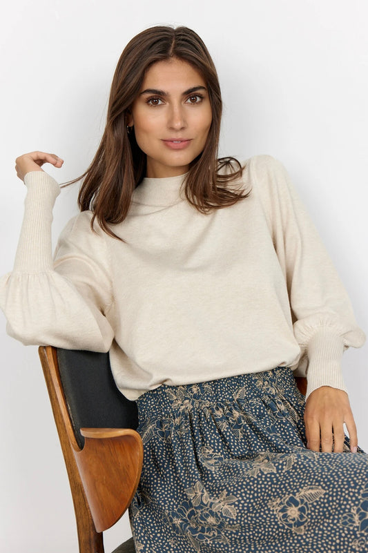 A woman with shoulder-length brown hair is seated on a wooden chair. She is wearing a cream melange Sweater Dollie 666 from SoyaConcept, featuring long puff sleeves and a standing collar, paired with a patterned skirt in shades of blue and beige. The background is plain white.