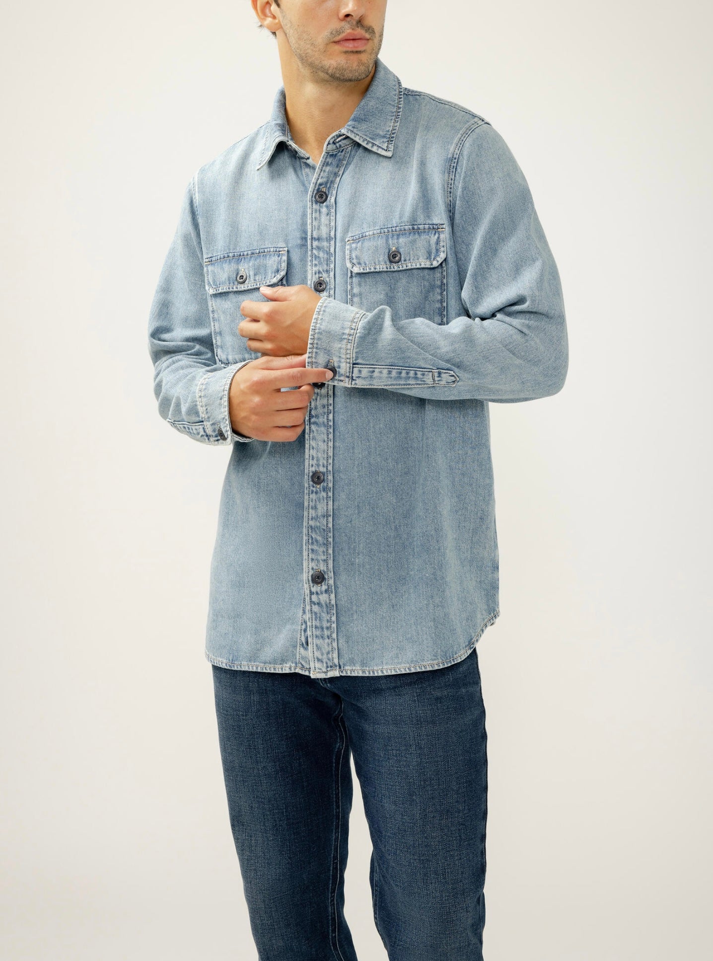 A man in a Silver Jeans Co. Denim Button-Up Shirt MJ0035LCL275 in medium indigo with chest pockets and a relaxed fit, paired with dark blue jeans, stands against a plain light background adjusting his sleeve.