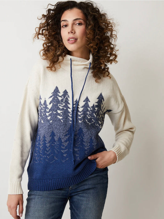 Wearing the THALIA TREE ECO COTTON HIGH NECK/FUNNEL PULLOVER SWEATER 87209 by Parkhurst, a person with curly hair stands comfortably with one hand in their pocket, showcasing the cozy cream and blue design. Made from eco-friendly fabric, the sweater's funnel neck features drawstrings that add charm to their casual jeans outfit against a plain background.