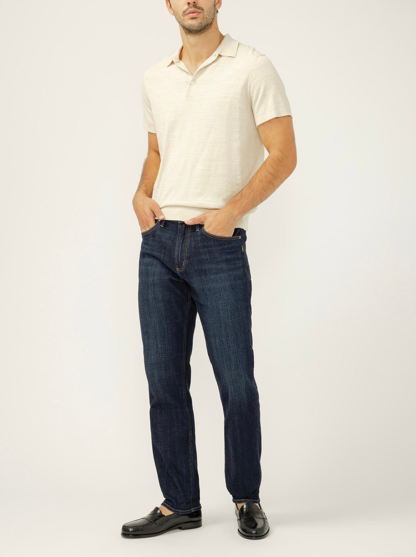 A man stands against a plain background with his hands in his pockets, wearing a cream short-sleeve polo, black loafers, and Silver Jeans Co. Machray Classic Fit Slim Straight Leg Jeans M63412LDS499 in dark indigo.