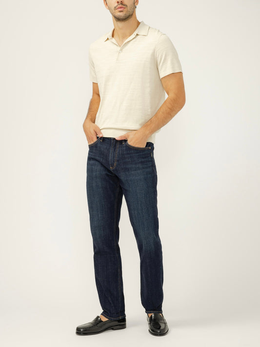 A man stands against a plain background with his hands in his pockets, wearing a cream short-sleeve polo, black loafers, and Silver Jeans Co. Machray Classic Fit Slim Straight Leg Jeans M63412LDS499 in dark indigo.