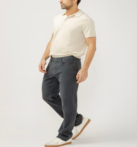 A man in a light beige polo, Silver Jeans Co. Grayson Classic Fit Straight Leg Knit Flex Jeans (M23425KTS668), and white sneakers stands against a plain light background, facing slightly sideways with one leg forward and arms relaxed.