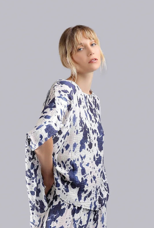 A woman with blonde hair in a loose updo wears the Paolo Tricot Top LX50153B Navy, a loose-fitting white top with blue abstract patterns, standing against a plain light gray background.