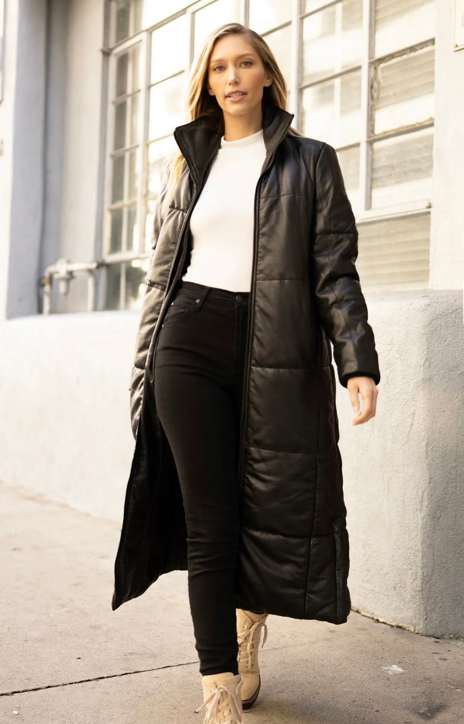 A person wearing the Evanna CF Leather Jacket, Black-Mauritius by MAURITIUS LEATHER over a white turtleneck and black pants walks confidently along a sidewalk, braving the winter chill. They have long light brown hair and beige boots. The person is in front of a building with large windows, the jacket providing extra warmth against the cold.