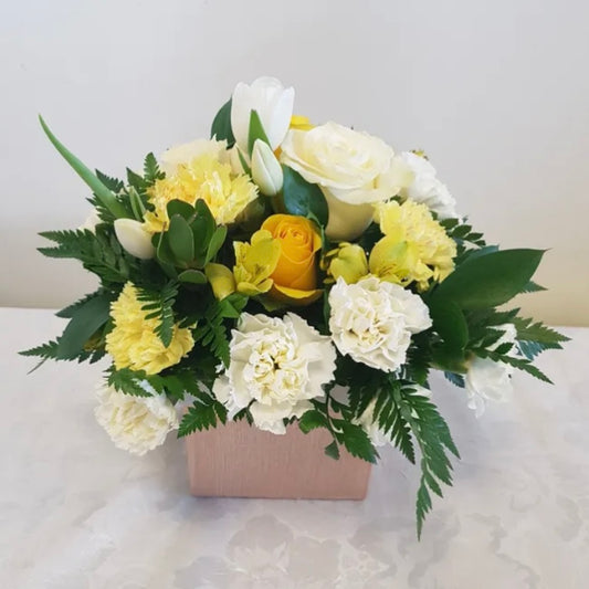 Presenting the "Sweet Whispers" bouquet from Posies Flowers & Fashion: a beautiful arrangement of white roses, yellow carnations, white tulips, and lush greenery in a square wooden container. Photographed against a plain white background, this elegant display is perfect for brightening any space. Check availability today!