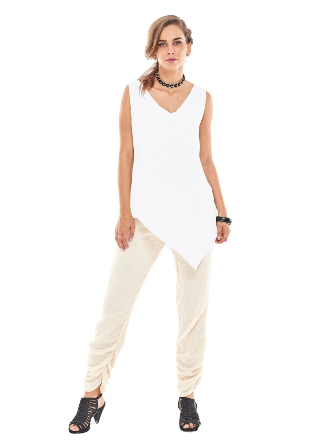 A confident woman stands against a white background wearing the ALLIE BLOUSE SNOW, an asymmetrical V-neck sleeveless top from Oh My Gauze. She pairs it with beige crinkled pants and black open-toed ankle boots, accessorizing with a black choker necklace and black bracelets. One hand rests confidently on her hip.