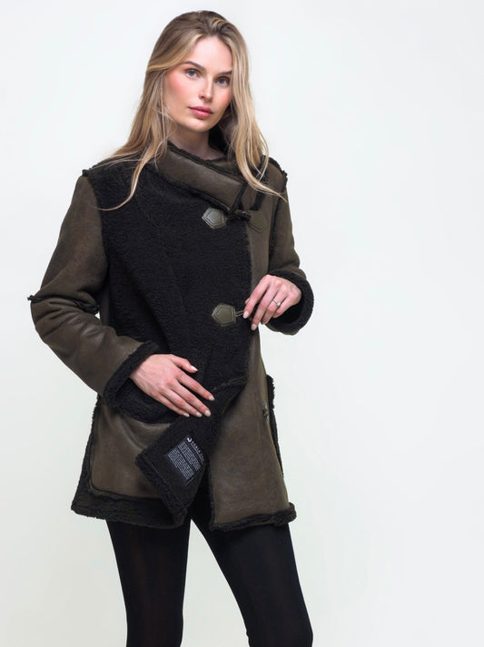 A woman with long, blonde hair is wearing a stylish Noble Coat - CHAKEN in dark green and black with asymmetrical buttons and black leggings. She stands against a plain light background, posing with one hand in her coat pocket and the other resting on the edge of her coat.