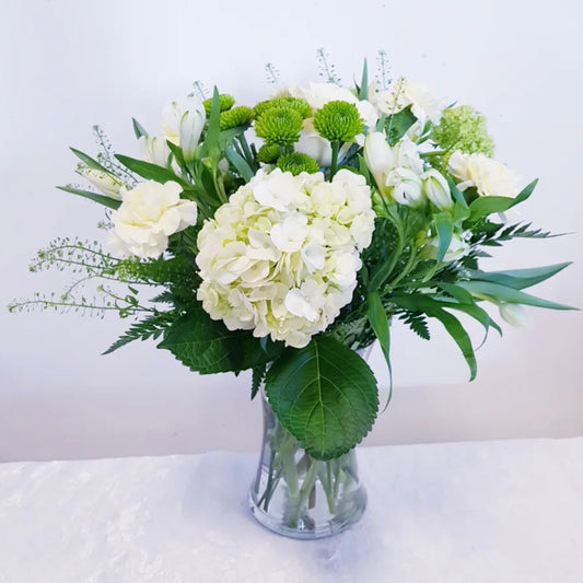 The "White Dove" arrangement by Posies Flowers & Fashion features a clear glass vase filled with an exquisite blend of white and green flowers, including hydrangeas, love-symbolizing roses, and green button poms. The bouquet is beautifully accented with lush greenery. The purity of the display is further enhanced by placing the vase on a light surface against a plain white background.