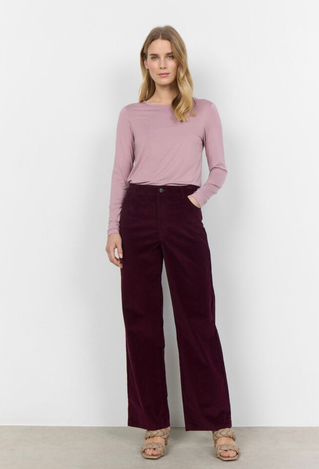 A person wearing a light pink long-sleeve shirt and SoyaConcept's SC-IVALOU 3-C dark purple velvet organic cotton straight-leg pants stands on a light-colored floor. Their feet are partially visible, showing flat, beige braided sandals. The background is plain and light-colored.