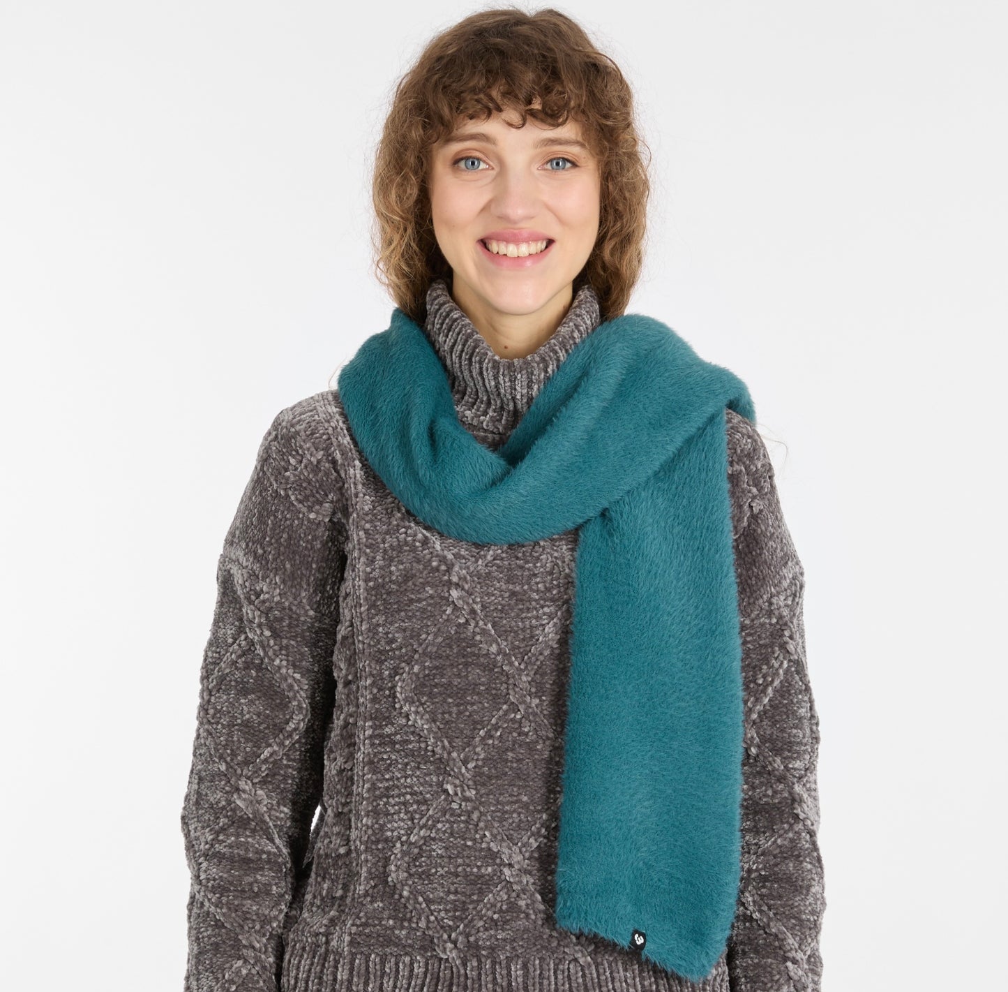 A person with curly brown hair smiles in a gray patterned sweater and the YANCY Scarf by RAGWEAR (ocean blue), standing against a plain white background.