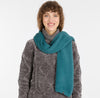 A person with curly brown hair smiles in a gray patterned sweater and the YANCY Scarf by RAGWEAR (ocean blue), standing against a plain white background.