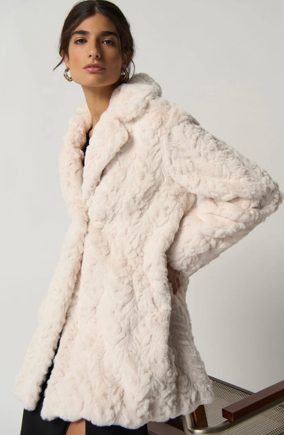 A woman with dark hair, wearing a white JOSEPH RIBKOFF COAT 233942 made from recycled polyester and gold hoop earrings, is leaning against a chair. She has a confident expression, and the background is plain light gray.