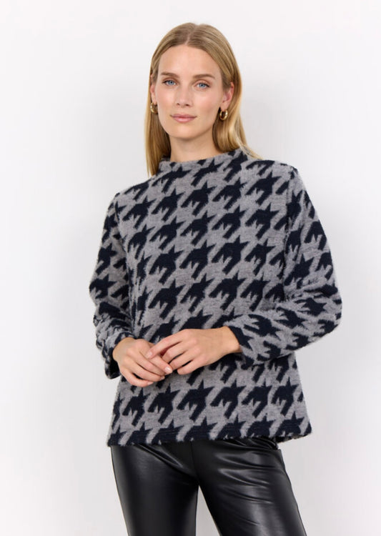 A person with long hair is wearing the SC-IRIT 1 Top from SoyaConcept, a high-neck, long-sleeve sweater adorned with a large black and grey houndstooth pattern. Over it, they are clad in a loose fit feminine waistcoat made of cotton blend. They also have on fitted black pants and are standing against a plain white background, looking straight at the camera.