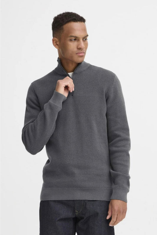 A man stands slightly turned, holding the collar of his dark gray BHBAAL HALF-ZIP KNIT blend -20720124 sweater from BLEND, paired with dark jeans against a plain white background.