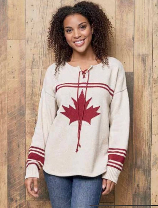 With curly hair and a smile, a person wears the Parkhurst CANADIANA ECO COTTON "HOCKEY" PULLOVER SWEATER 87137, featuring a relaxed fit and decorated with a red maple leaf. Standing against a wooden wall in blue jeans, they perfectly showcase this stylish sweater.