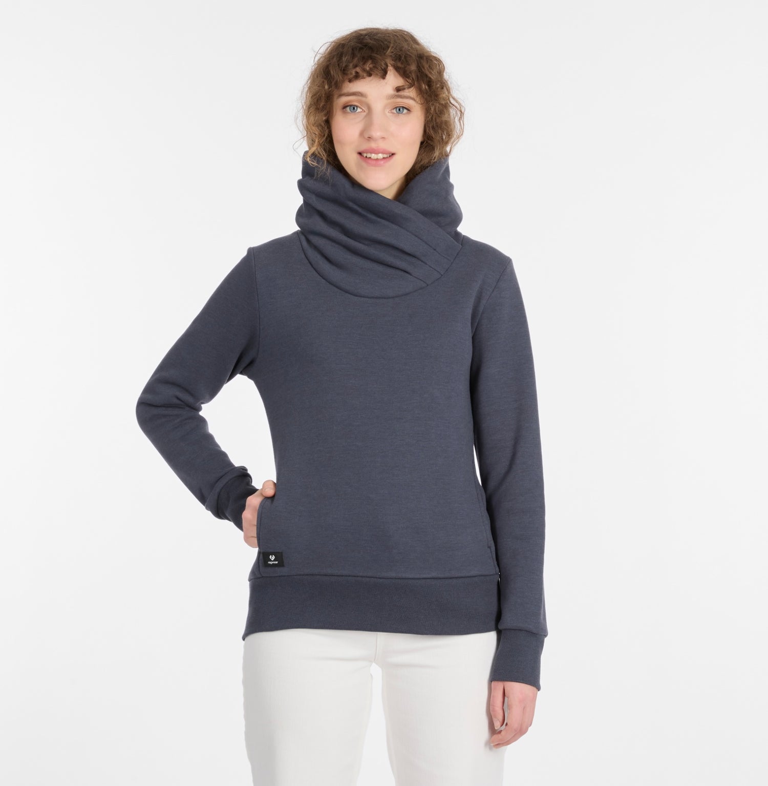 A woman with curly hair wears the ANABETH Women’s Sweatshirt by RAGWEAR in navy and white pants, standing against a plain white background, smiling with one hand in her pocket.